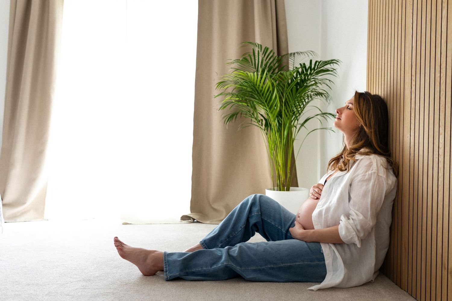 A pregnant woman in white lace lingerie rests peacefully on white bedding.
