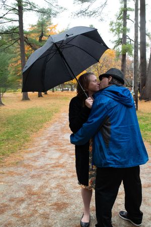 Two people share a black umbrella while walking on a wet autumn path through a park on a rainy day.