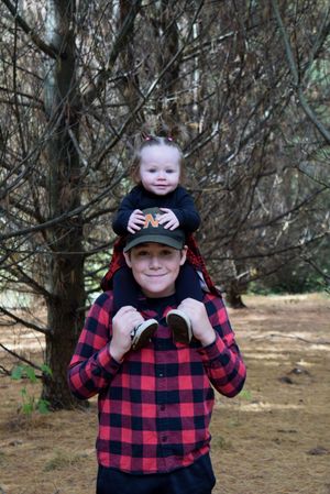 A young person in a red plaid flannel and baseball cap smiles while standing among trees in a forest setting.