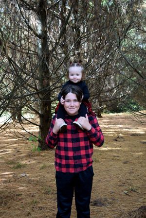 A child wearing a red and black buffalo plaid shirt and black pants poses playfully in a wooded outdoor setting.