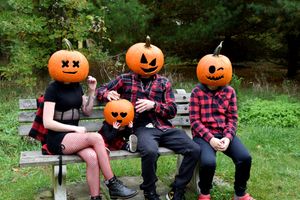 Four people sit outdoors on a bench wearing plaid shirts and holding carved Halloween pumpkins in their laps.