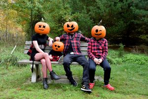 Three pumpkin-headed figures in plaid shirts and jeans sit casually on a wooden bench in an autumn garden setting.