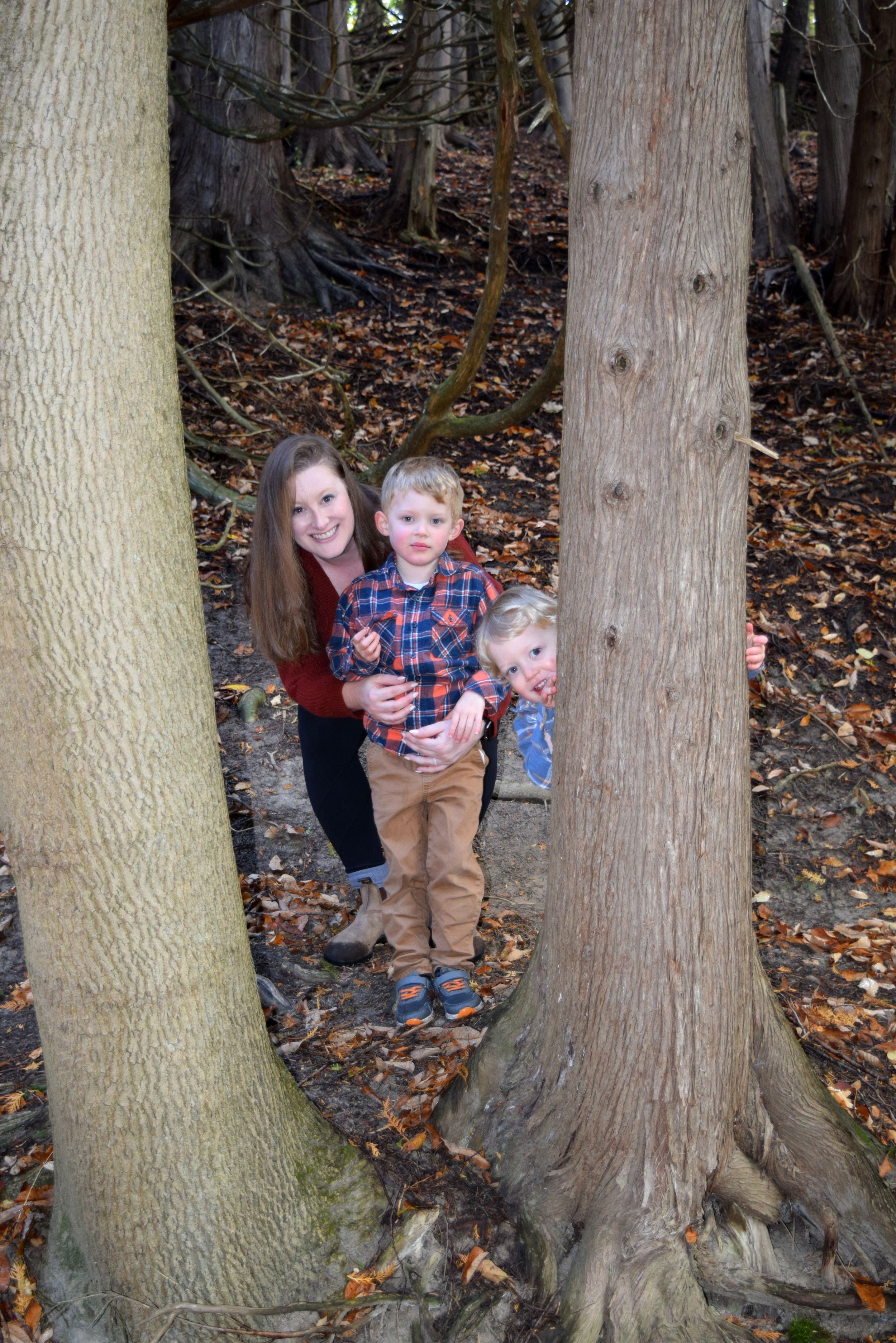 Children pose playfully between large tree trunks covered in autumn leaves in a forest setting.
