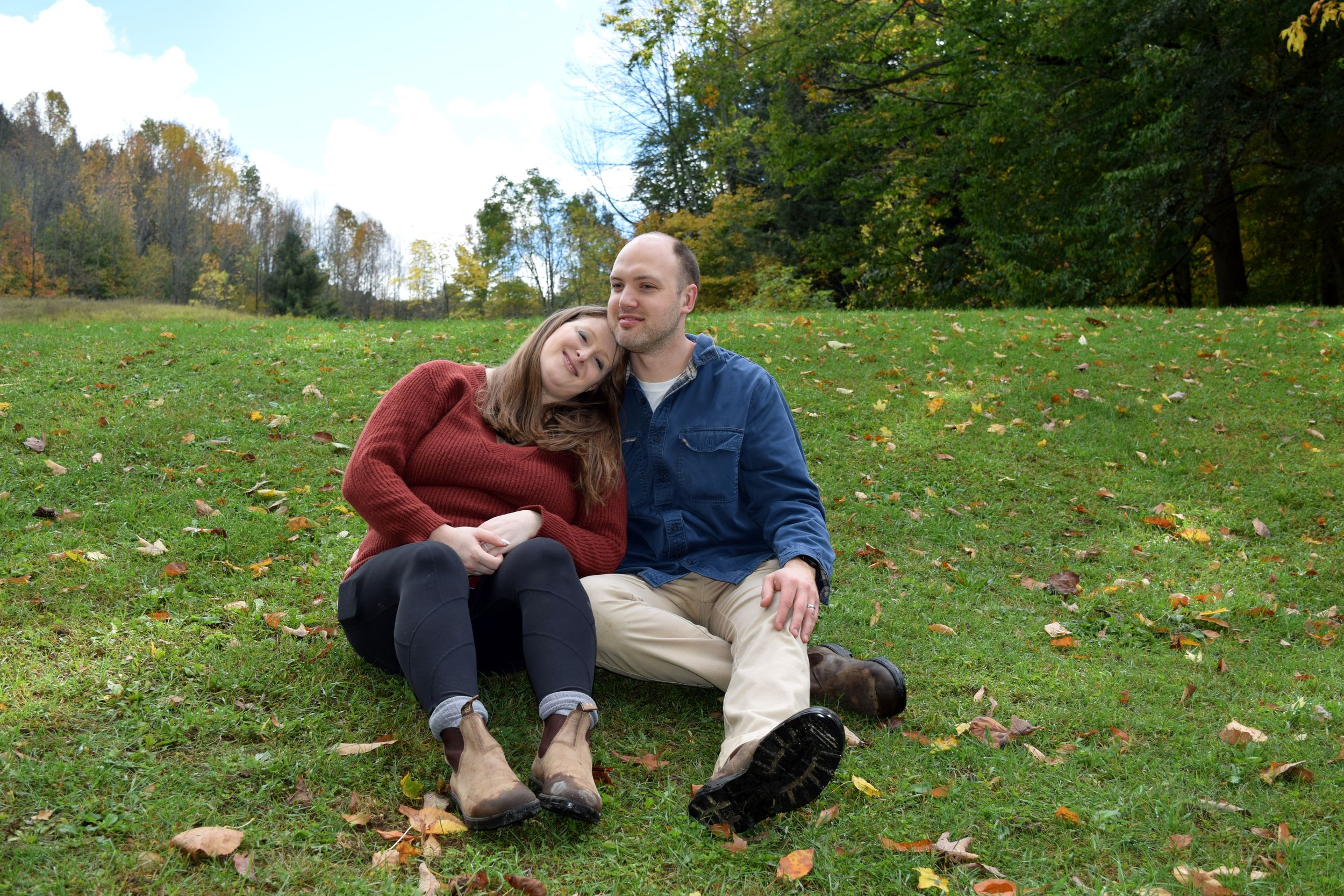 A couple sits together on grass covered with fallen autumn leaves in a park setting.