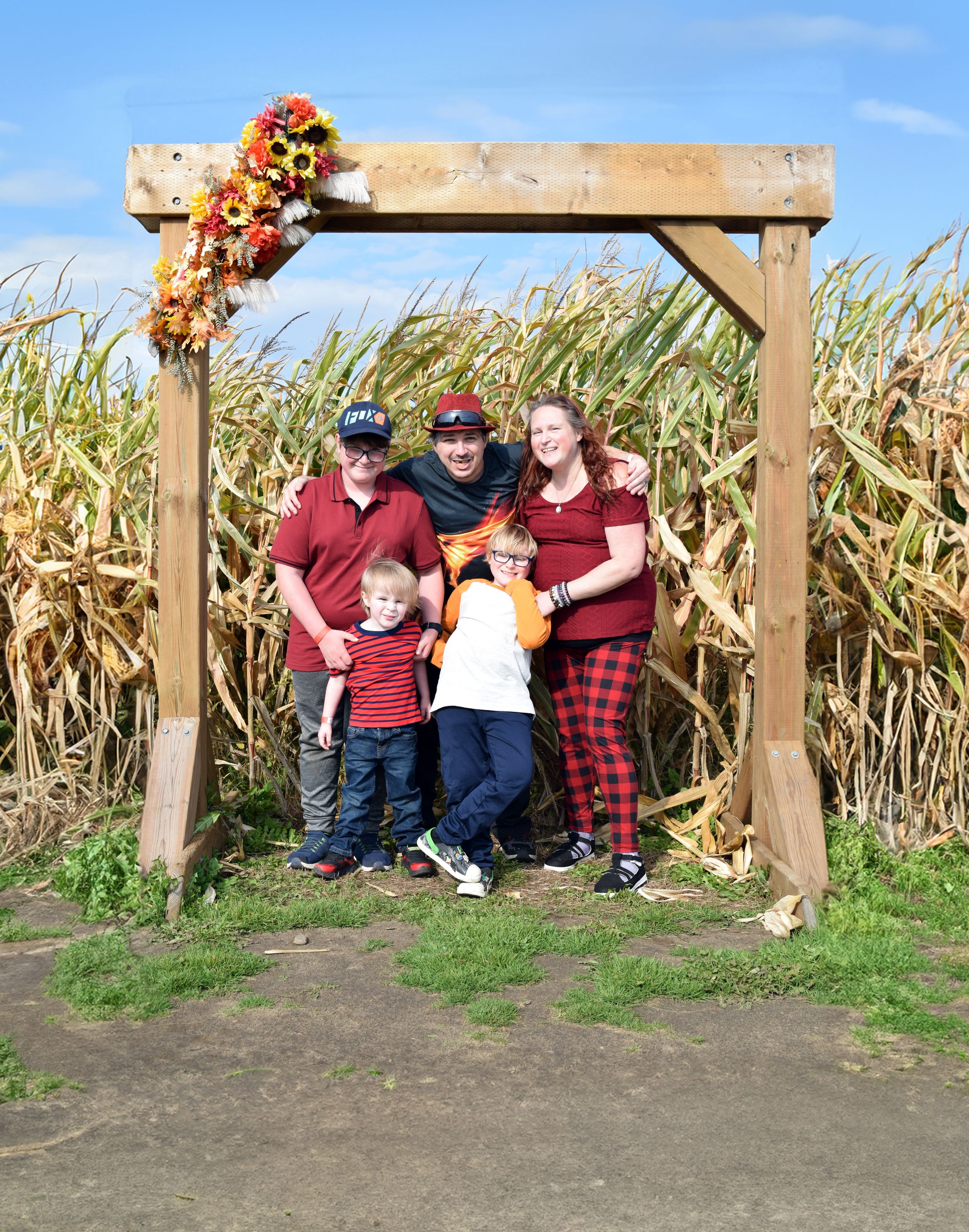 A group poses for photos at a wooden archway entrance to a corn maze on a sunny autumn day.