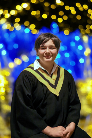 Graduate wearing black and gold academic regalia smiles against a bokeh background of blue and yellow lights.
