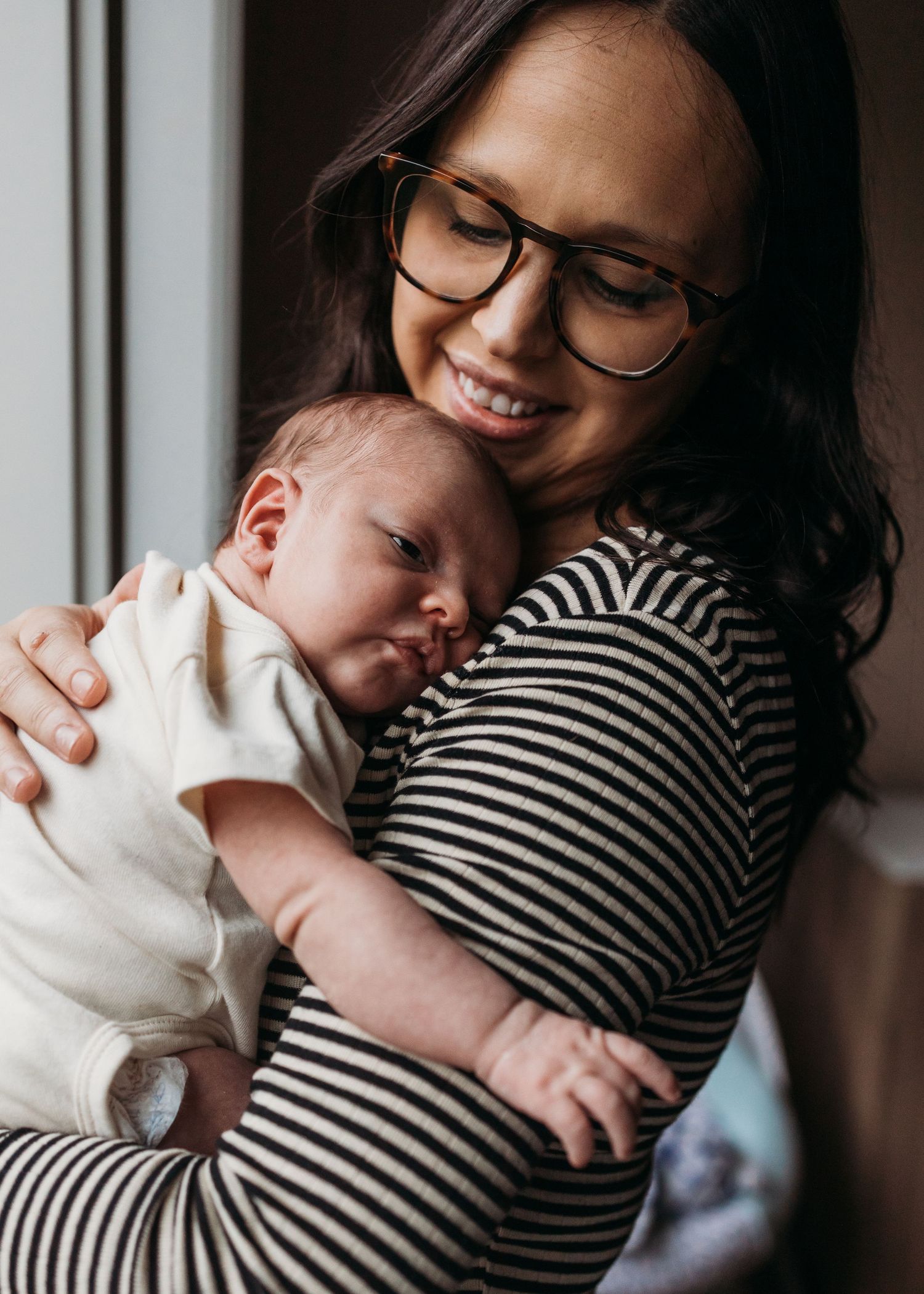 A parent wearing a striped shirt holds a sleeping baby in white clothing against a window with vertical blinds.