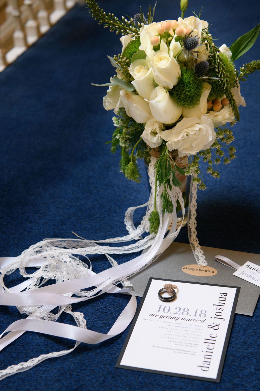 Wedding bouquet of white roses and greenery beside an invitation for Danielle and Joshua dated 10.28.18.