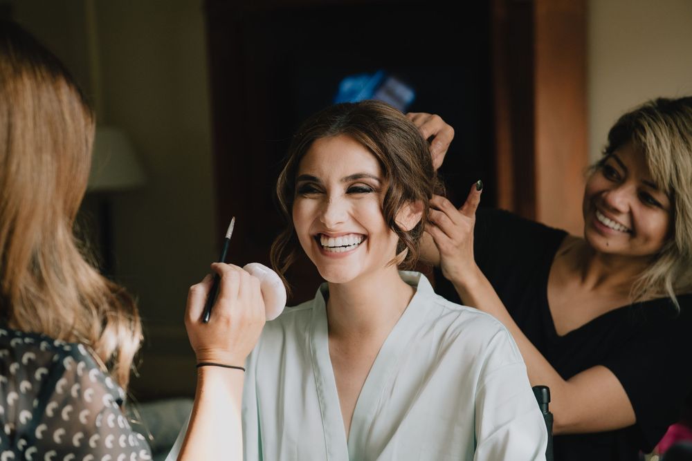 Bride smiling joyfully while getting makeup and hair done by two stylists in a white robe before her wedding.