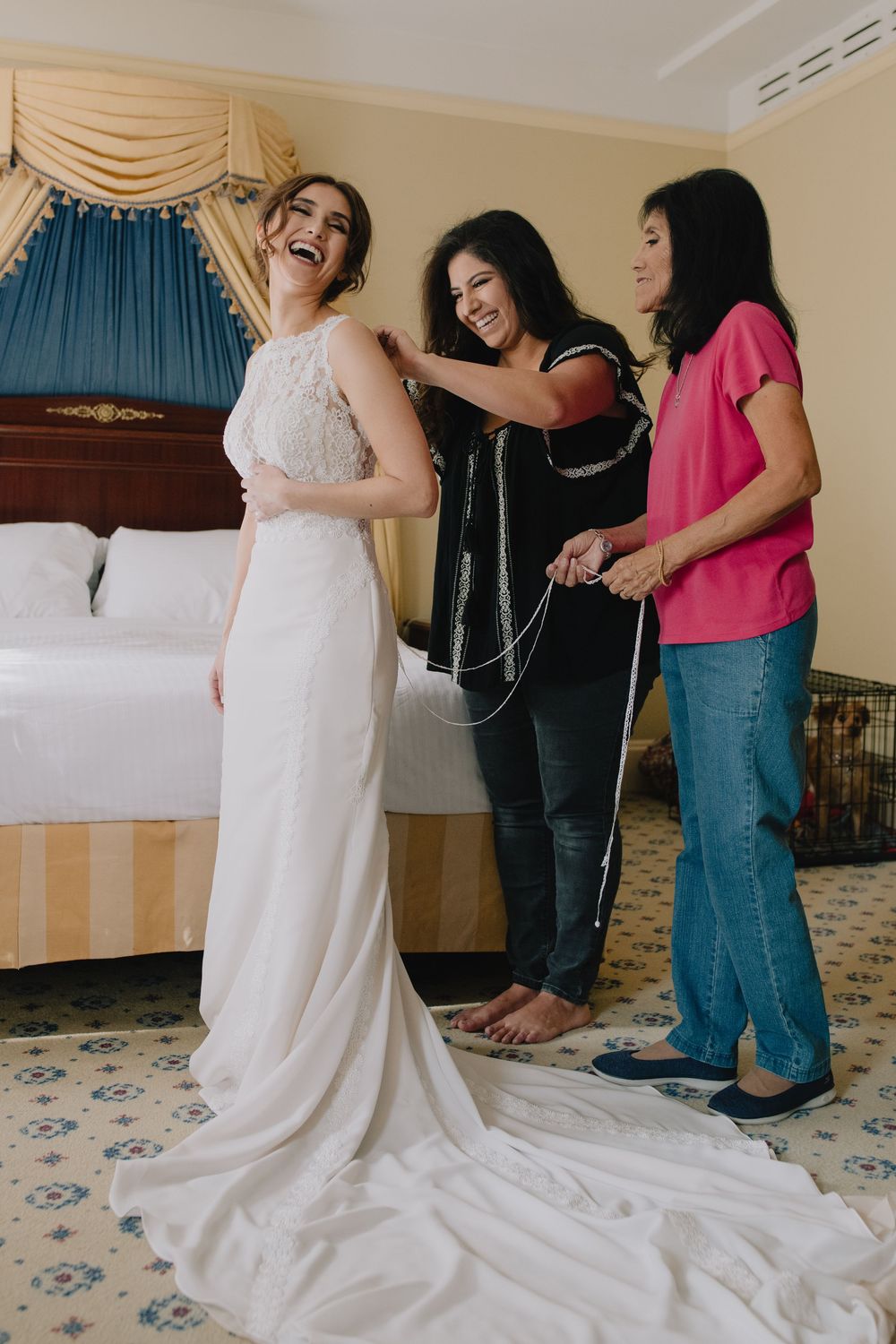 Bride laughing as two women help her into her elegant white wedding gown in a hotel room.