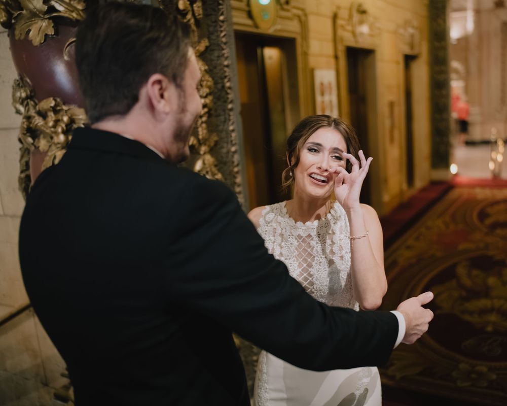 Emotional bride wiping tears during first look with groom in ornate wedding venue.