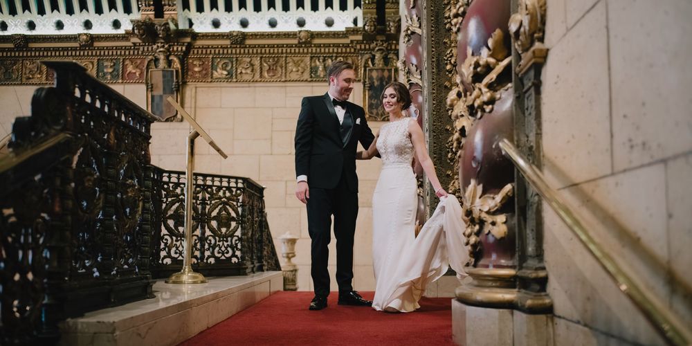 Bride and groom descend ornate staircase with red carpet in elegant historic venue during wedding ceremony.