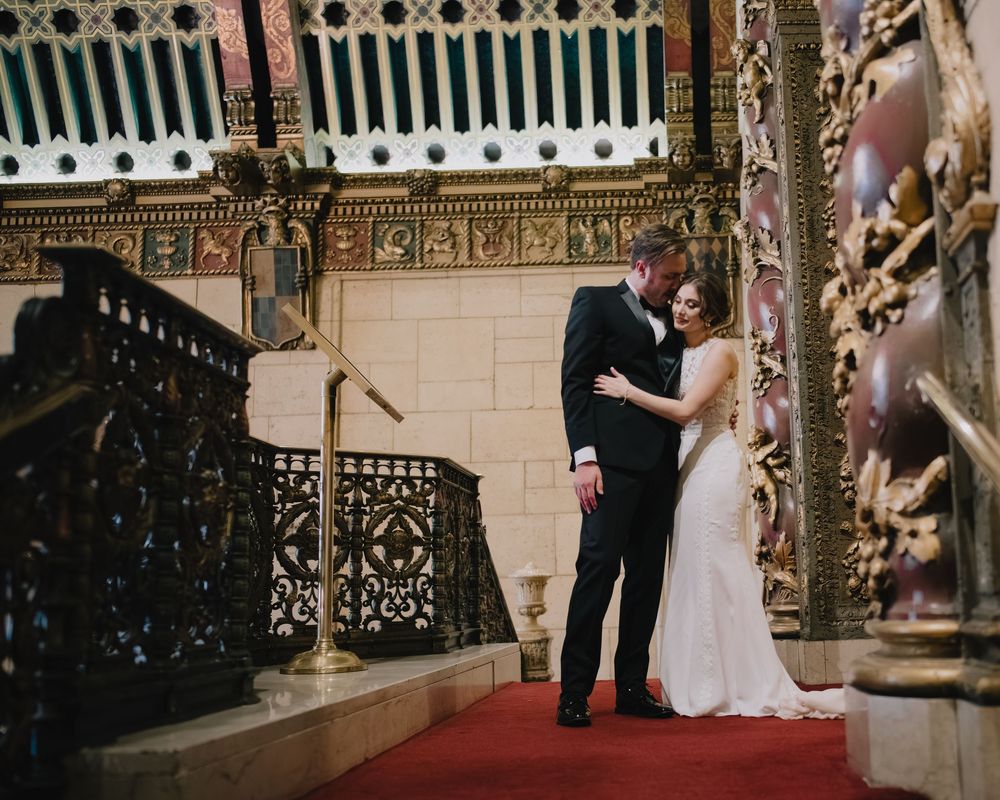 Bride and groom share a romantic moment on ornate staircase with red carpet in elegant historic venue.