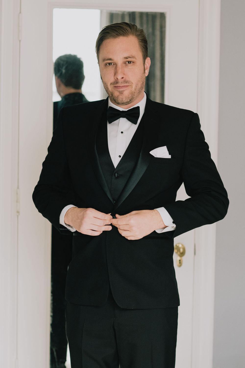 Groom in black tuxedo with bow tie buttoning jacket while preparing for wedding ceremony.