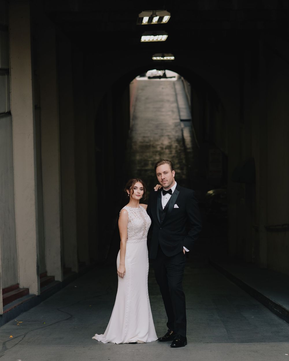 Bride and groom posing elegantly in archway, she in white gown, he in black tuxedo.