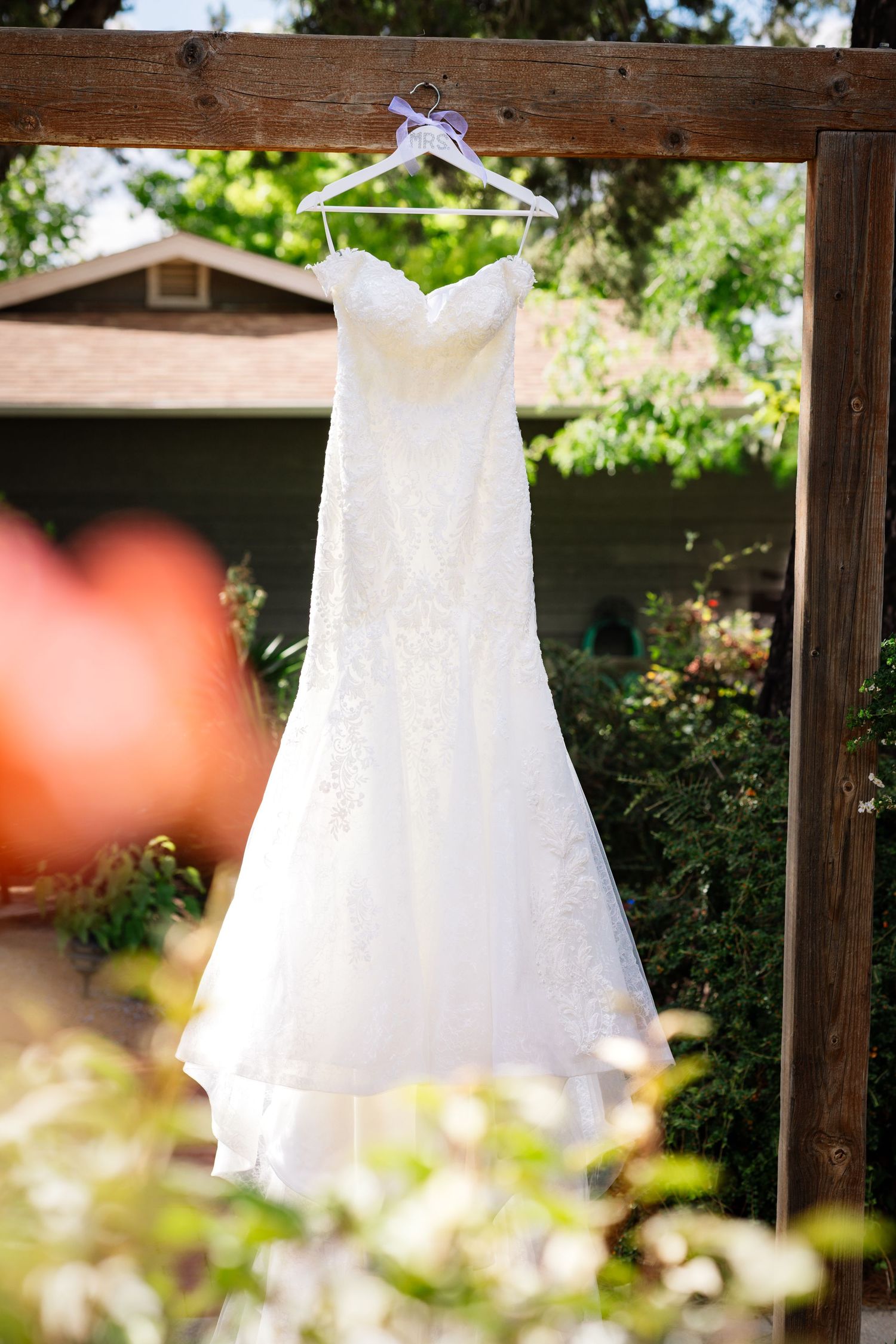 A white lace wedding dress hangs from a purple hanger against a wooden beam in a garden setting.