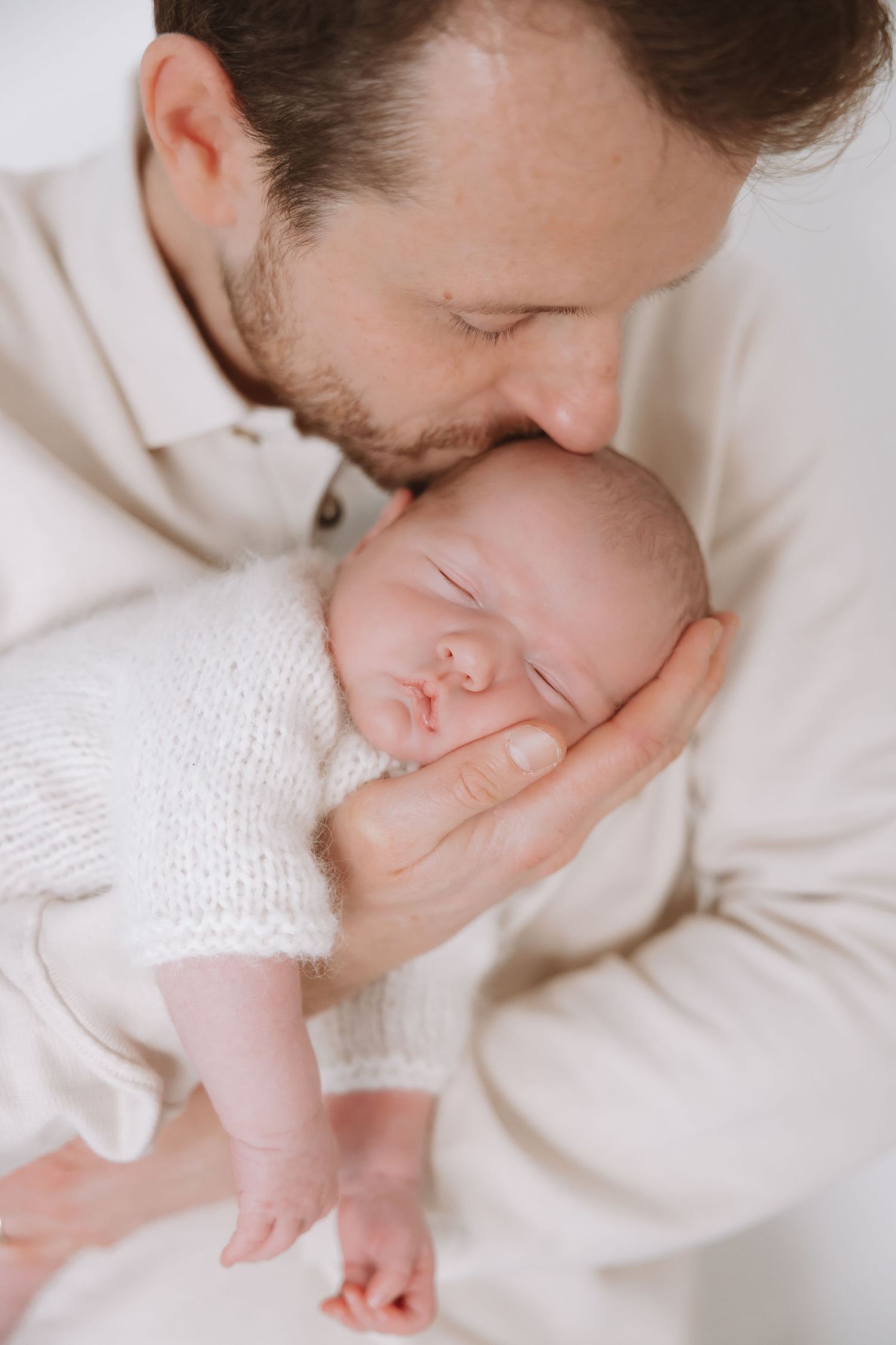 A tender moment between parent and newborn baby in soft white knit clothing against light background.