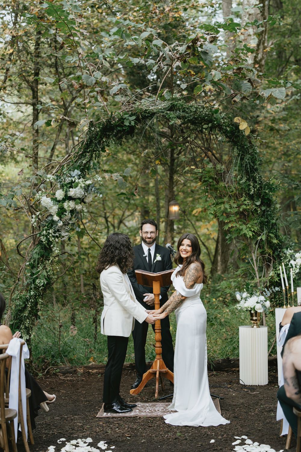 A romantic outdoor wedding ceremony takes place in a wooded setting with white decor and floral arrangements.