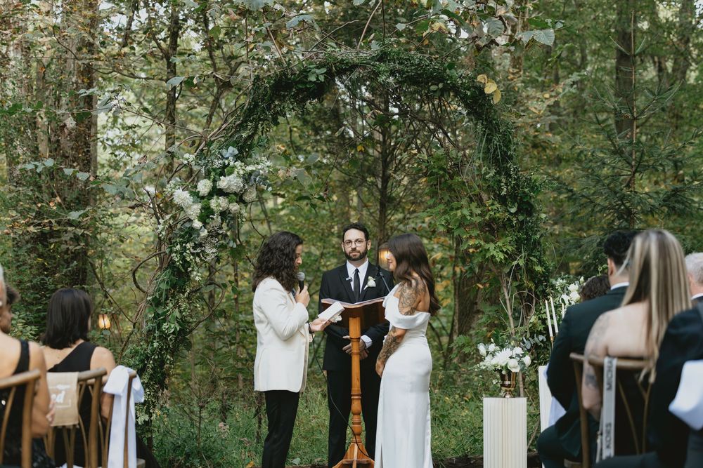 Outdoor wedding ceremony in a lush green forest setting with guests seated on wooden benches.