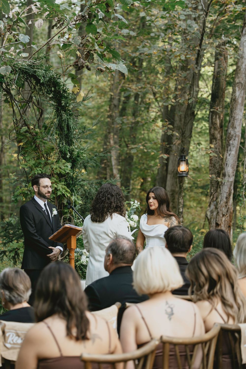 An intimate outdoor wedding ceremony takes place in a wooded setting with guests seated on wooden chairs among the trees.