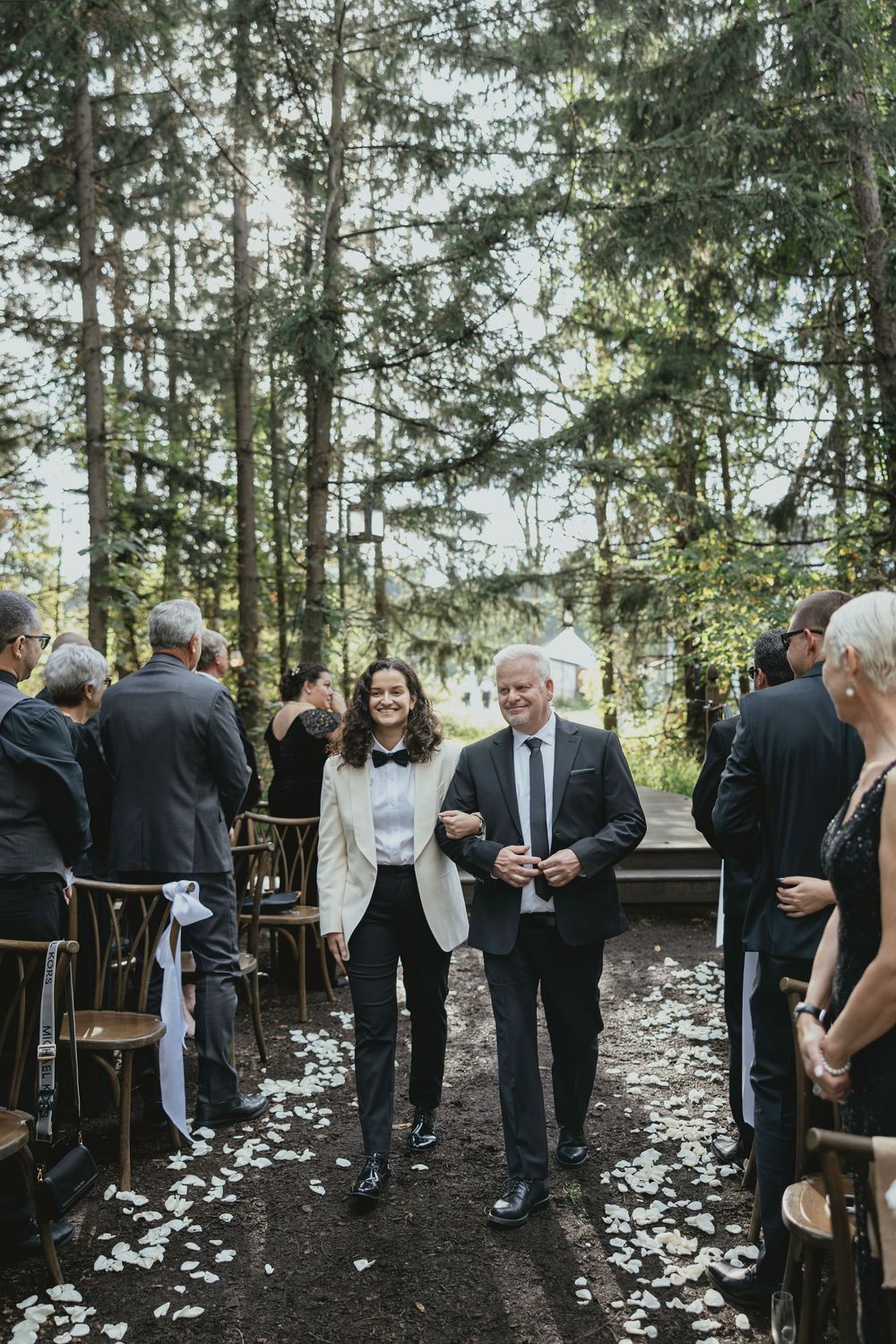 Wedding guests mingle outdoors on a wooded path covered in white flower petals during an intimate forest ceremony.