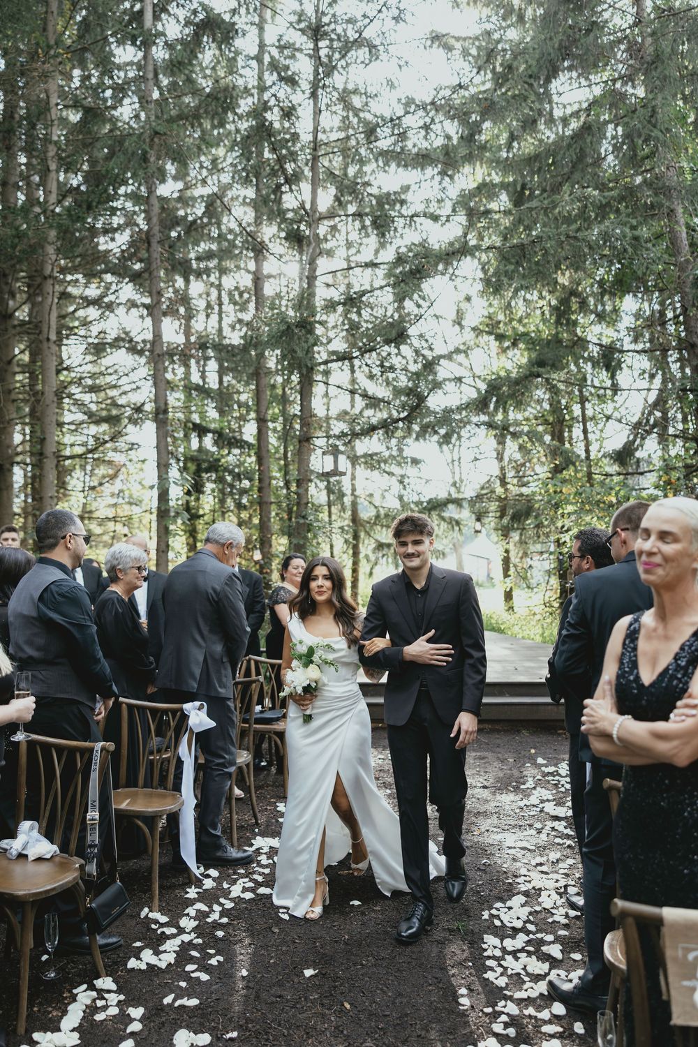 Wedding guests mingle in an outdoor forest setting with scattered white flower petals on the ground.
