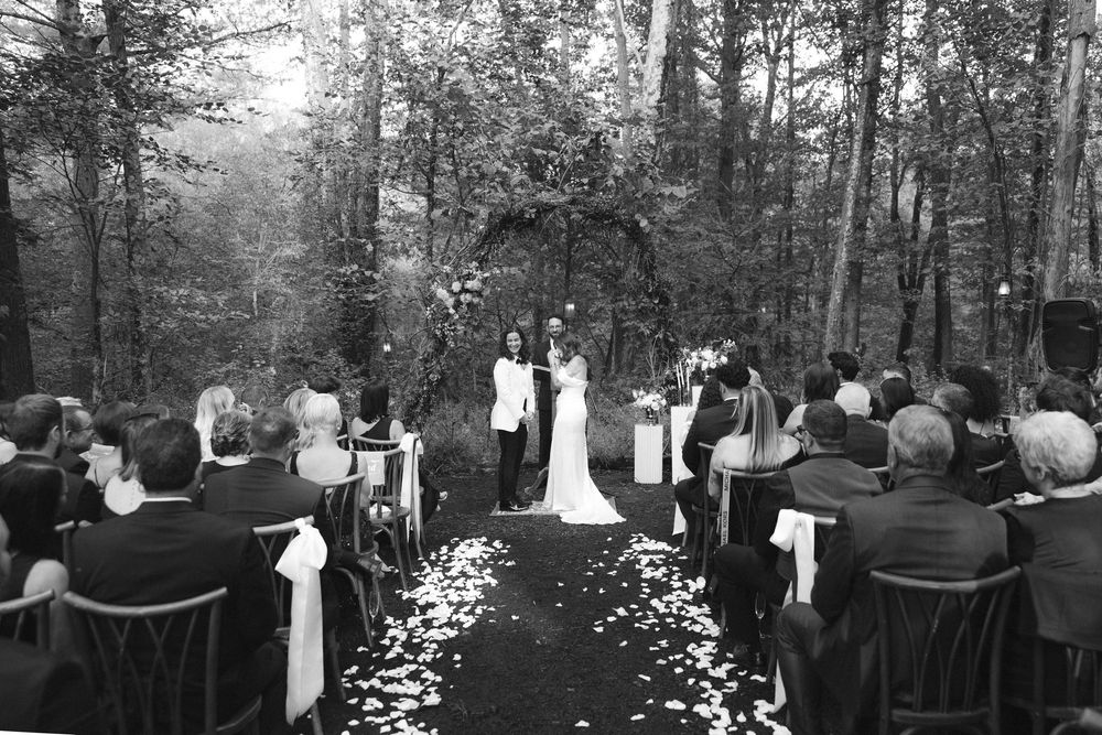 A panoramic black and white photograph of an outdoor wedding ceremony in a wooded forest setting with guests seated in rows.
