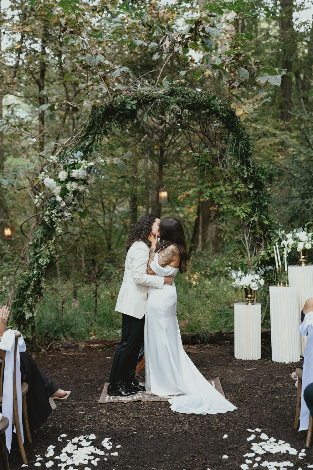 Two people in white dresses share an intimate moment during an outdoor ceremony with hanging lanterns and floral decor.