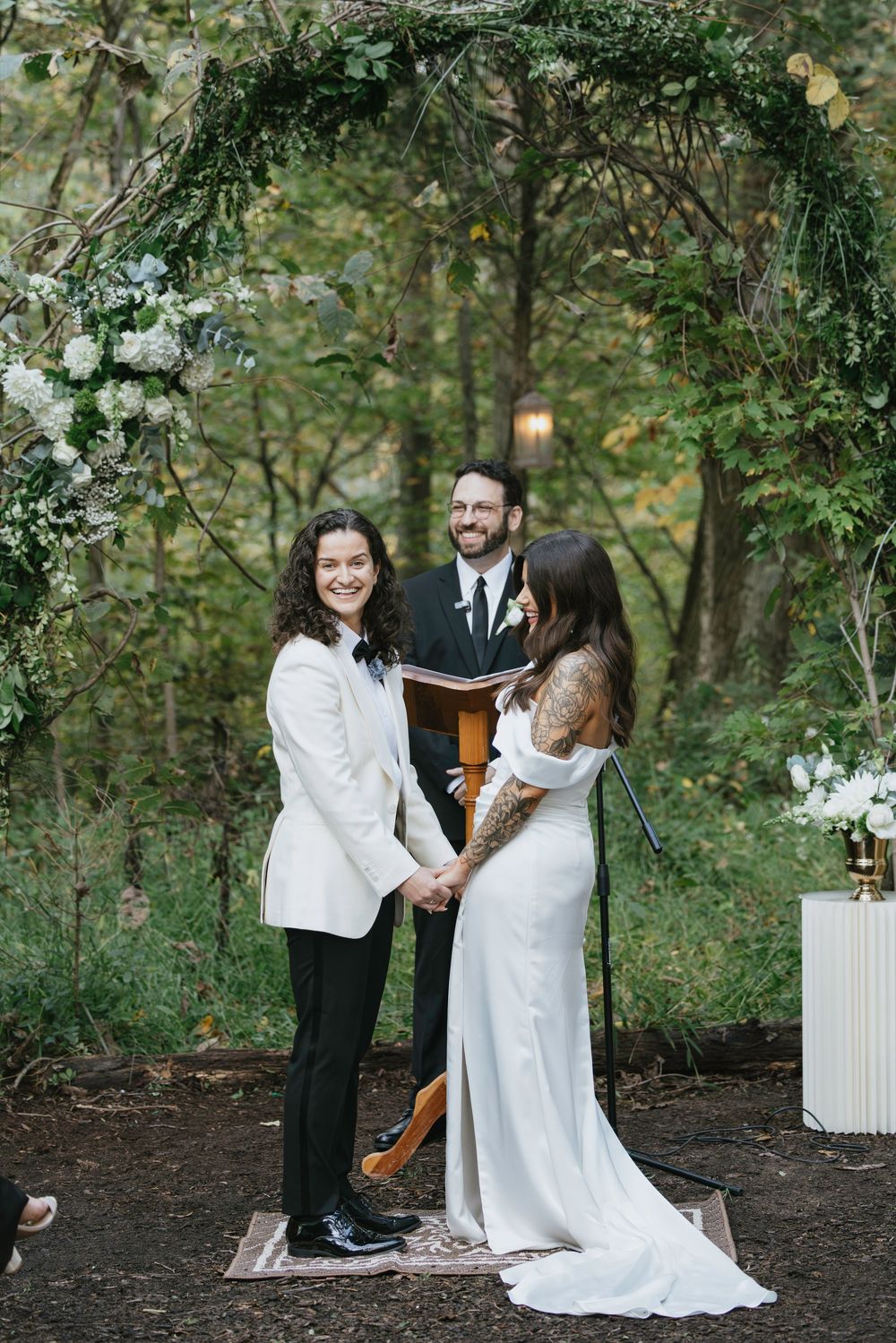 A couple exchanges vows under a lush green archway decorated with white flowers during an intimate forest wedding ceremony.