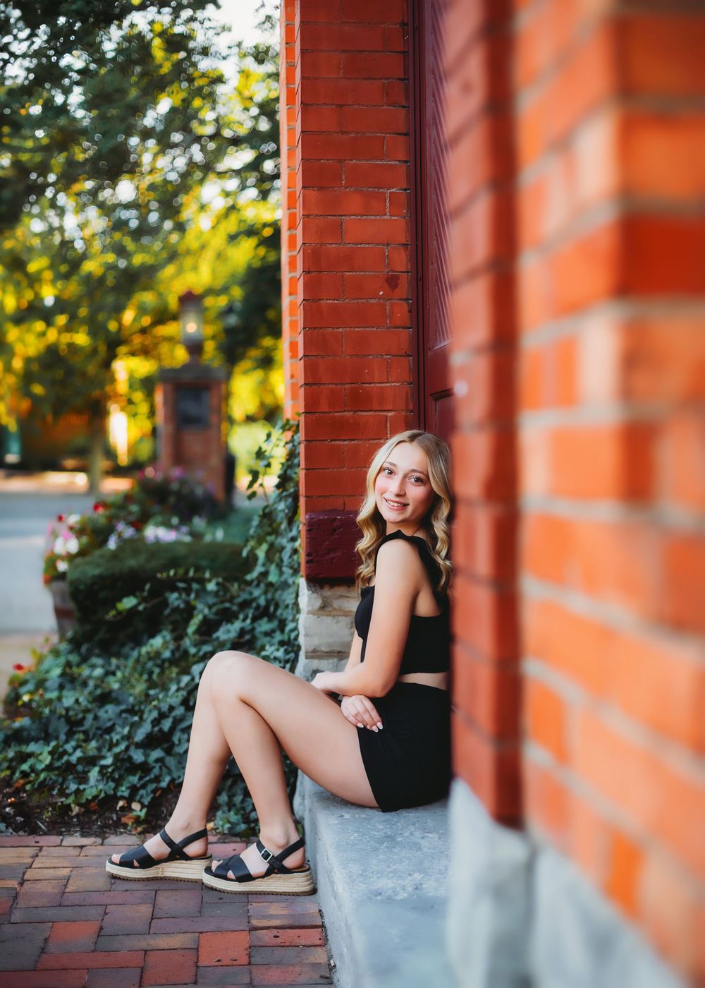 Portrait of someone sitting by red brick columns wearing a black outfit on a brick pathway.