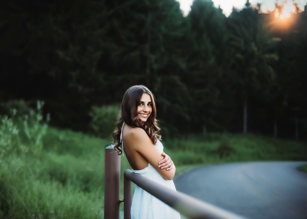 Woman in white dress poses against a guardrail at sunset with forest scenery in the background.