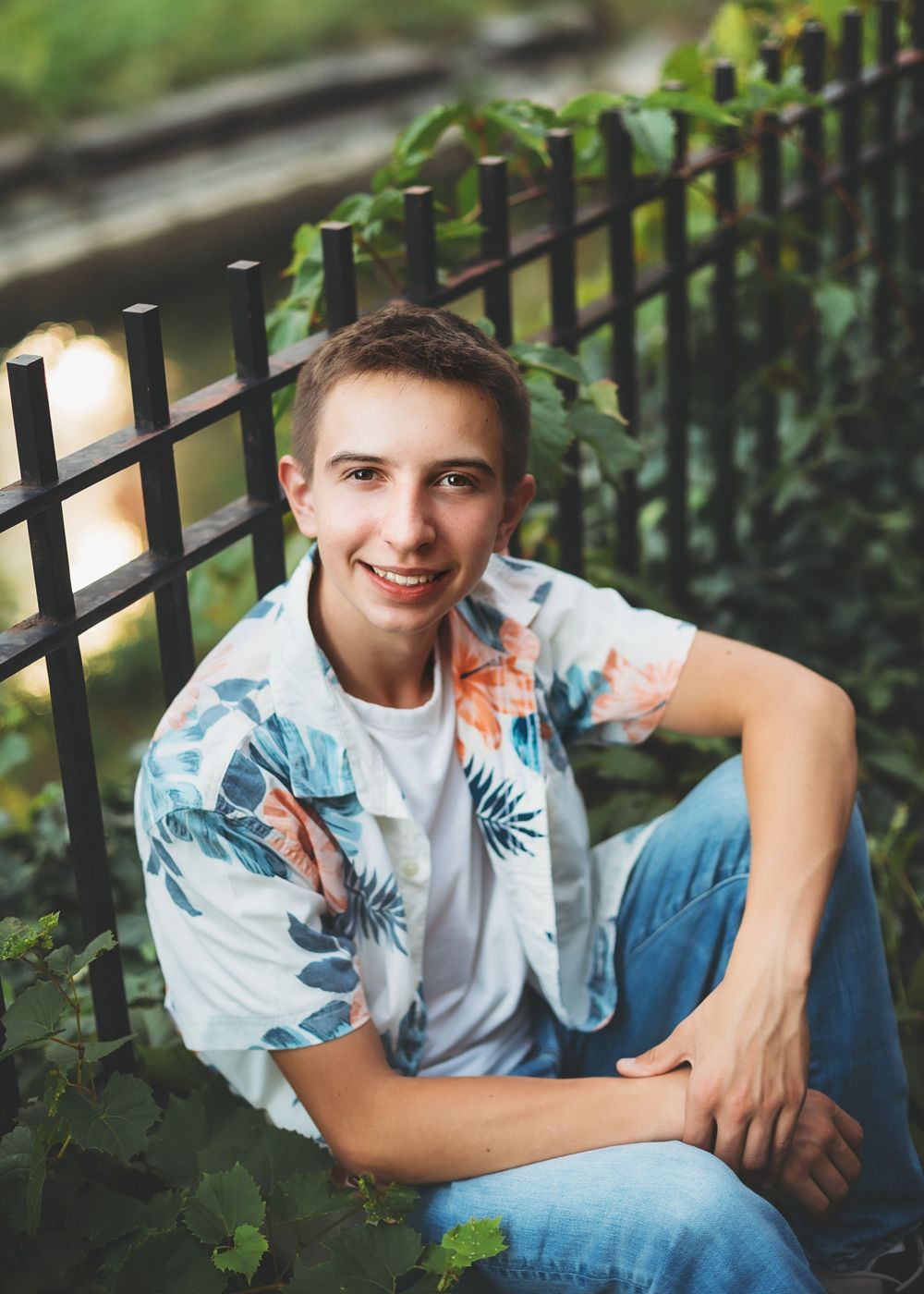 Young person in floral shirt and jeans sitting casually by an iron fence outdoors.
