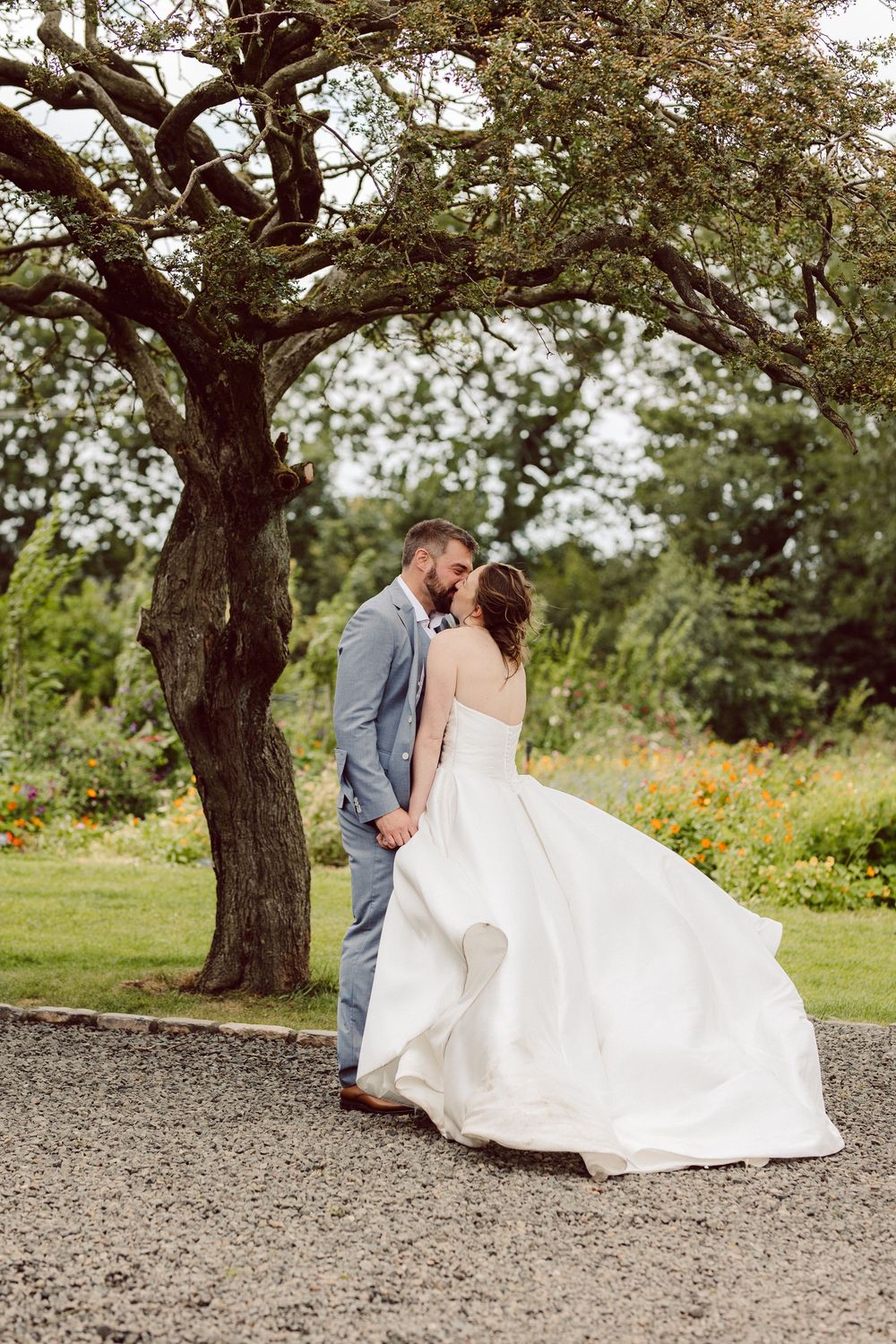 A couple in wedding attire walking together beneath a large tree in a rustic outdoor setting.