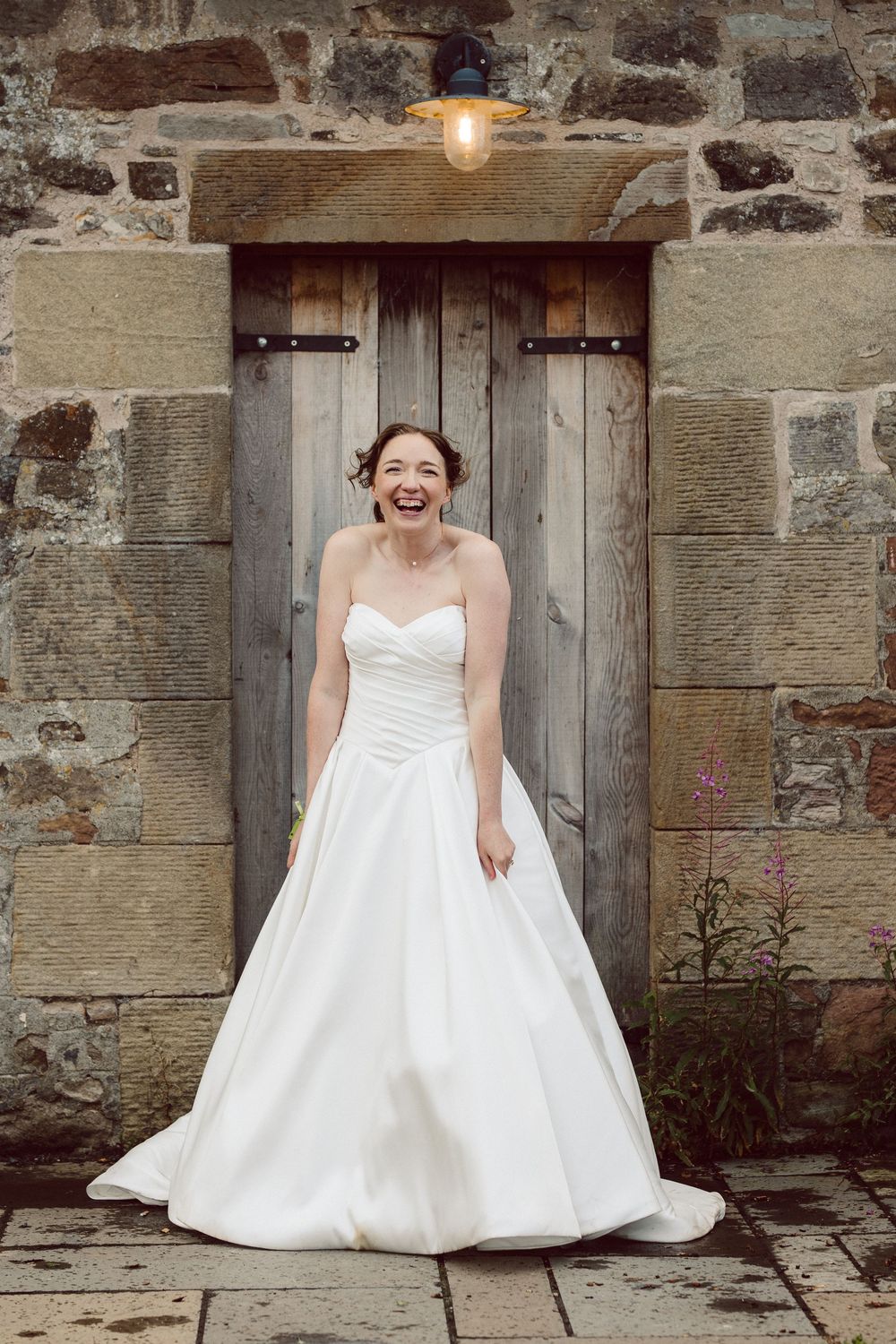A bride in a white wedding gown poses joyfully against a rustic stone wall and wooden door.