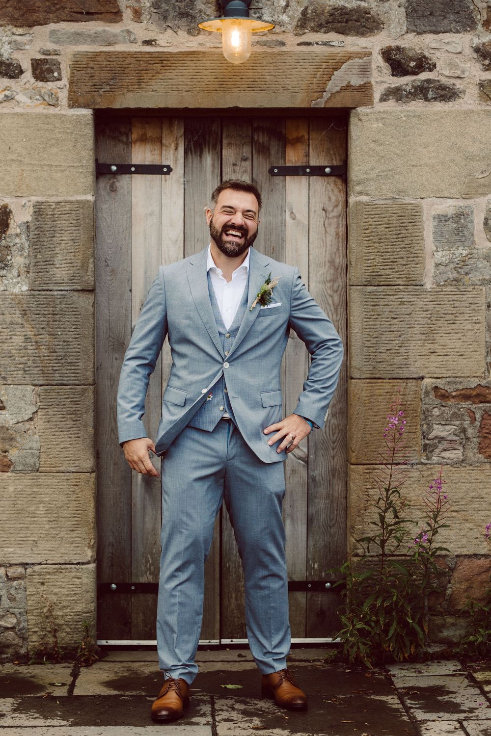 A person in a light blue suit poses joyfully against a rustic stone wall with a wooden door.