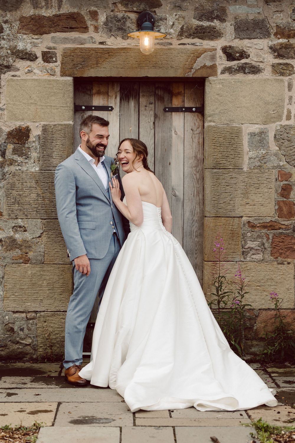 A couple in wedding attire poses in front of a rustic stone wall and wooden door with an overhead light.