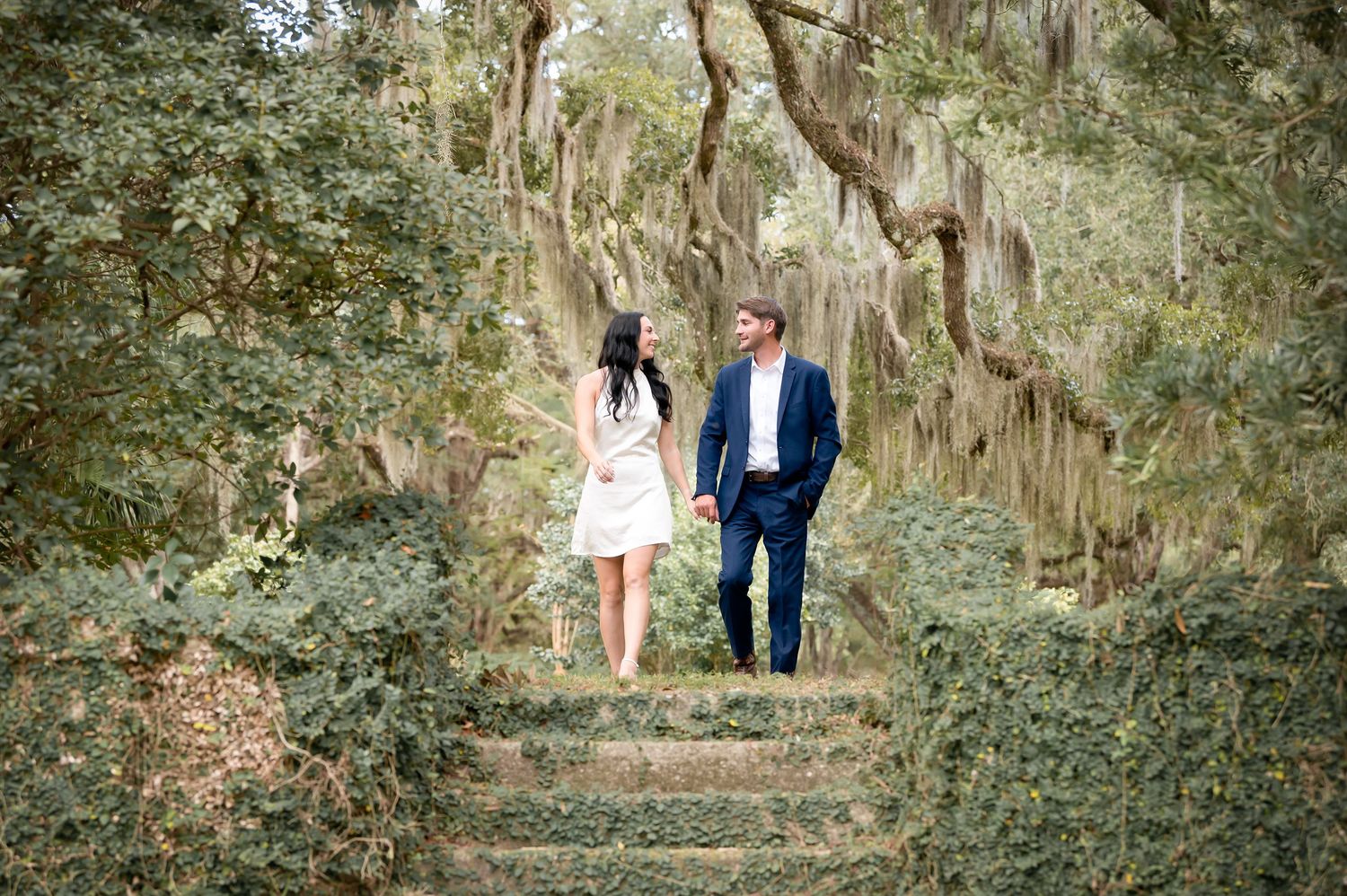 A couple walks hand in hand along a scenic path under moss-draped oak trees in a romantic outdoor setting.