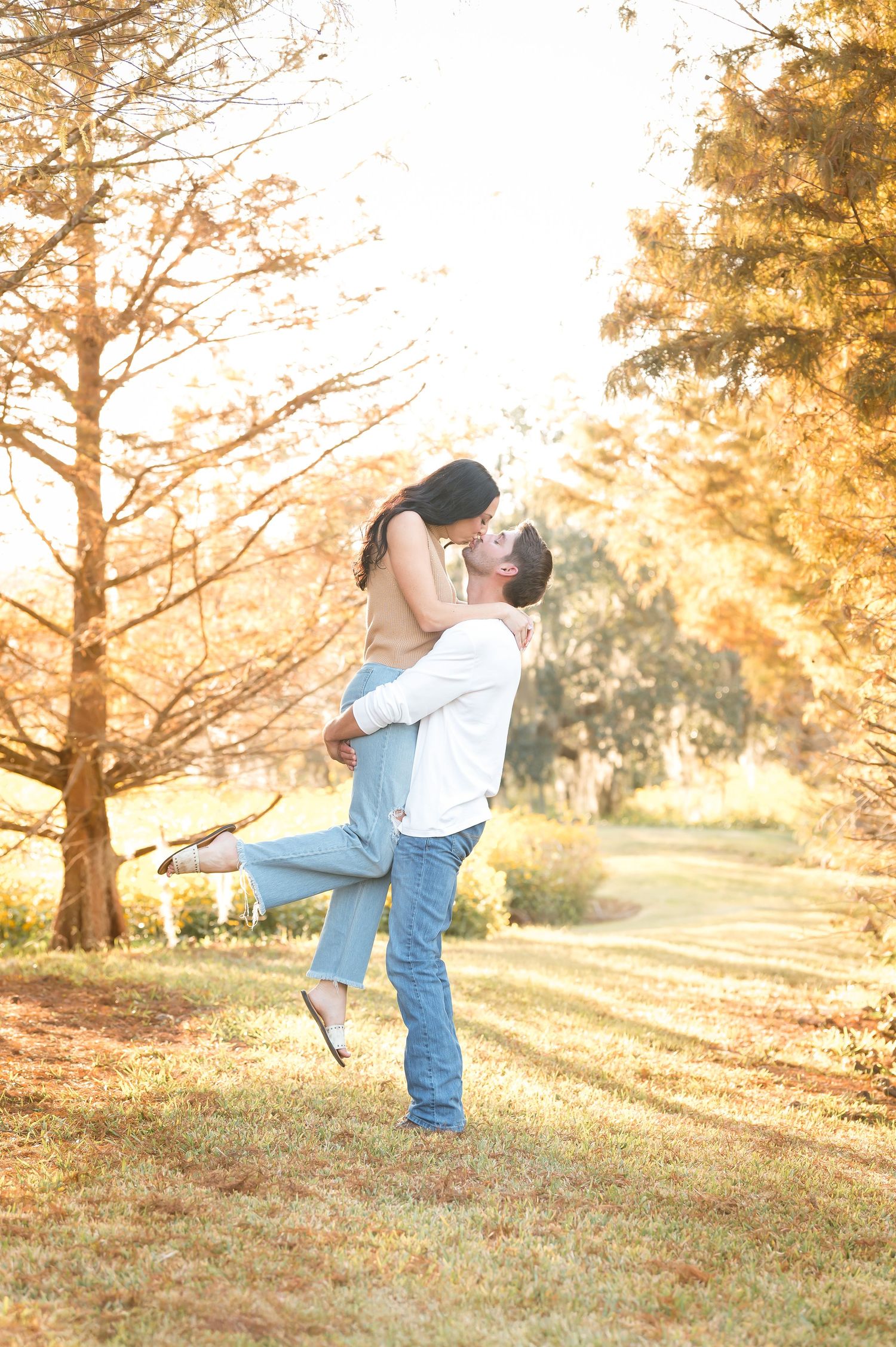 A romantic couple embraces in a sun-drenched meadow during golden hour with tall trees in the background.