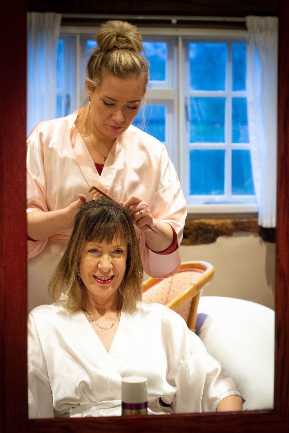 A hairstylist works on styling a client's hair in a salon with natural light coming through a window.