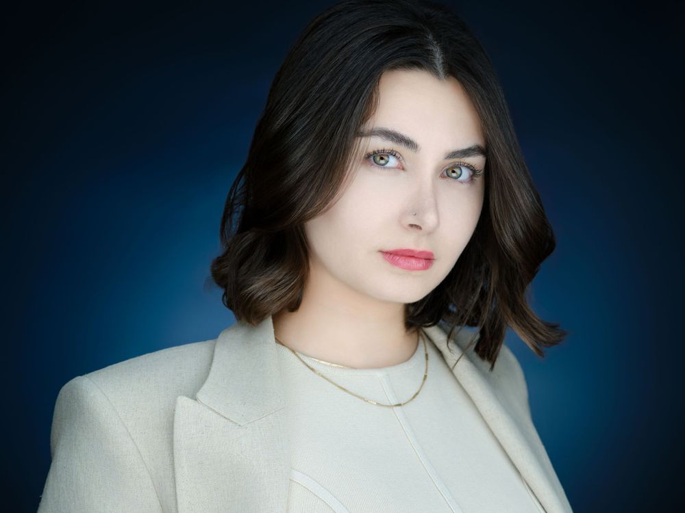 Professional corporate headshot of a young woman in white blazer with dark hair on dark blue background.