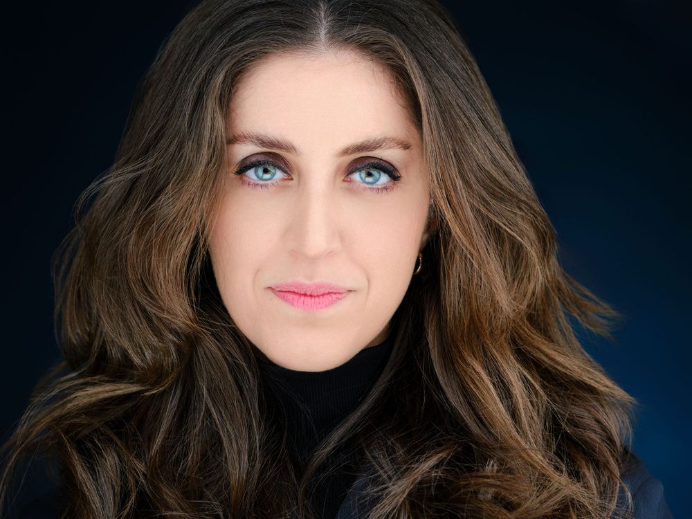 Professional corporate headshot of a woman with wavy brown hair and blue eyes on dark blue background.
