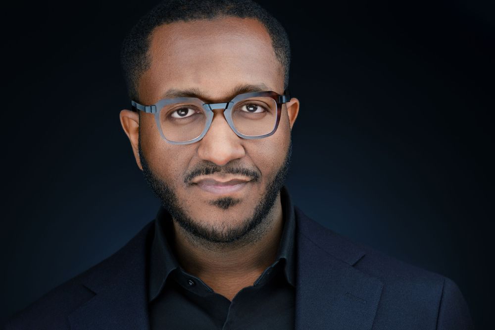Professional corporate headshot of a man wearing glasses and dark suit jacket on dark blue background.