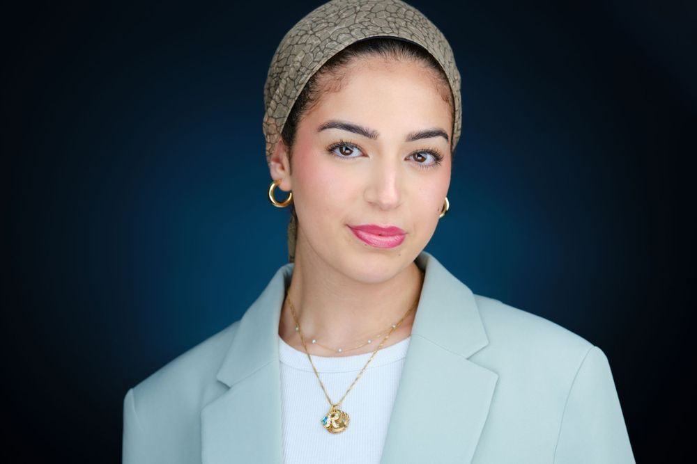 Professional corporate headshot of a young woman wearing a headscarf and light blue blazer on dark background.