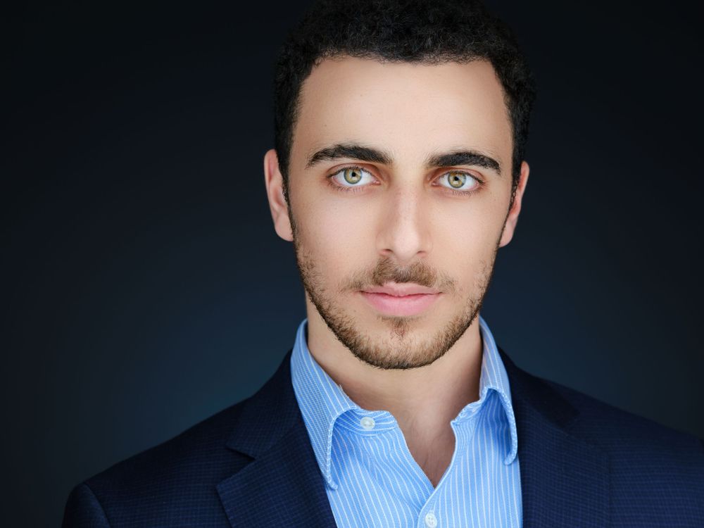 Professional corporate headshot of a young man in blue dress shirt and suit jacket on dark background.