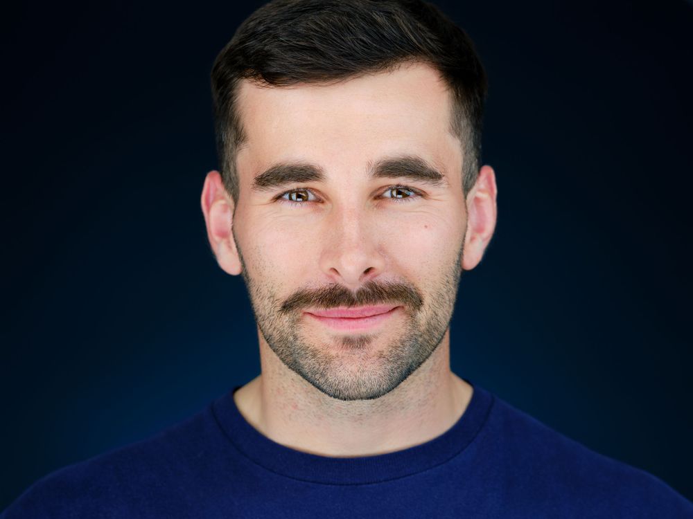 Professional corporate headshot of a smiling man with beard wearing a navy blue sweater on dark background.