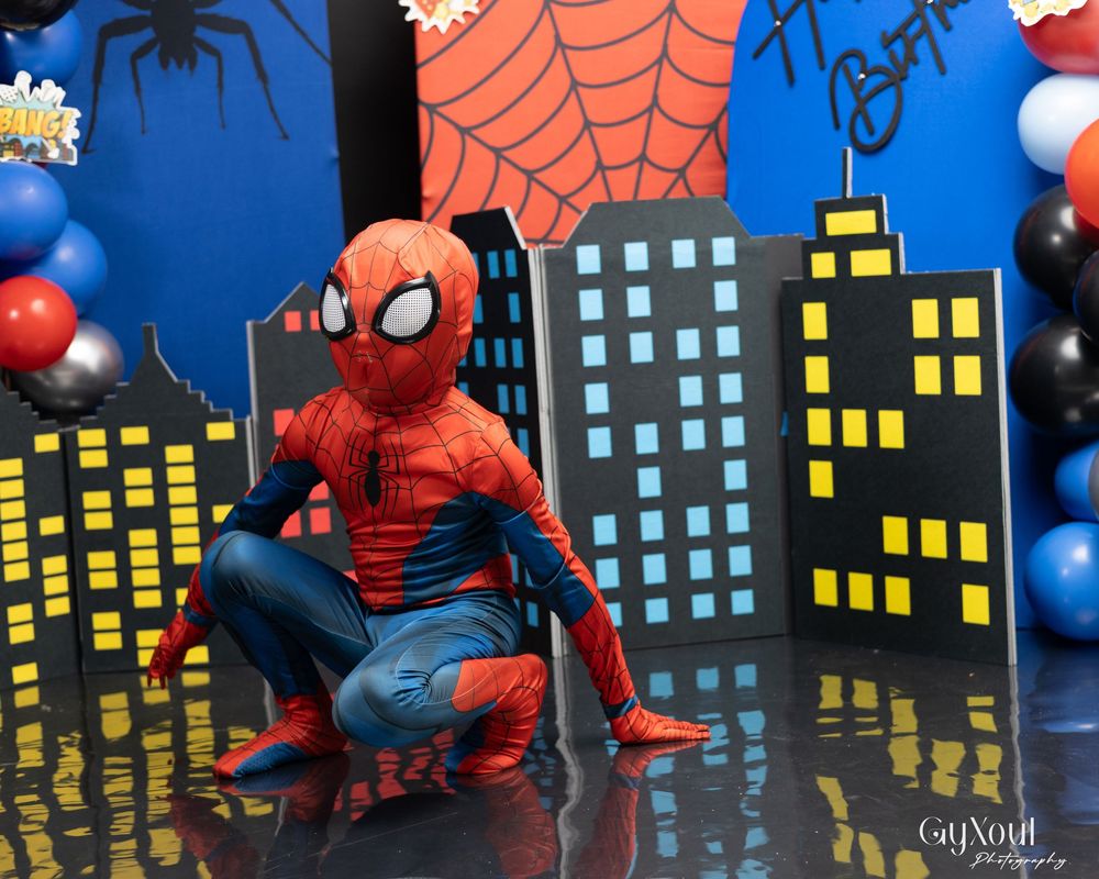 Spider-Man figure crouches against a cityscape backdrop with balloons and spider web decorations at a party setup.
