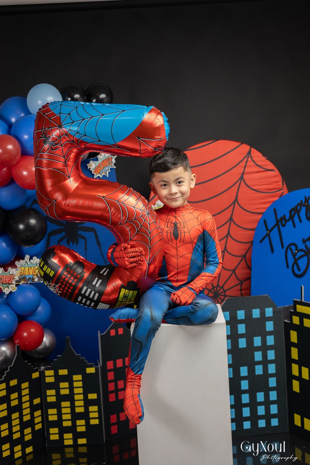 A Spider-Man themed birthday party backdrop features a cityscape silhouette with red and blue balloons against a white wall.