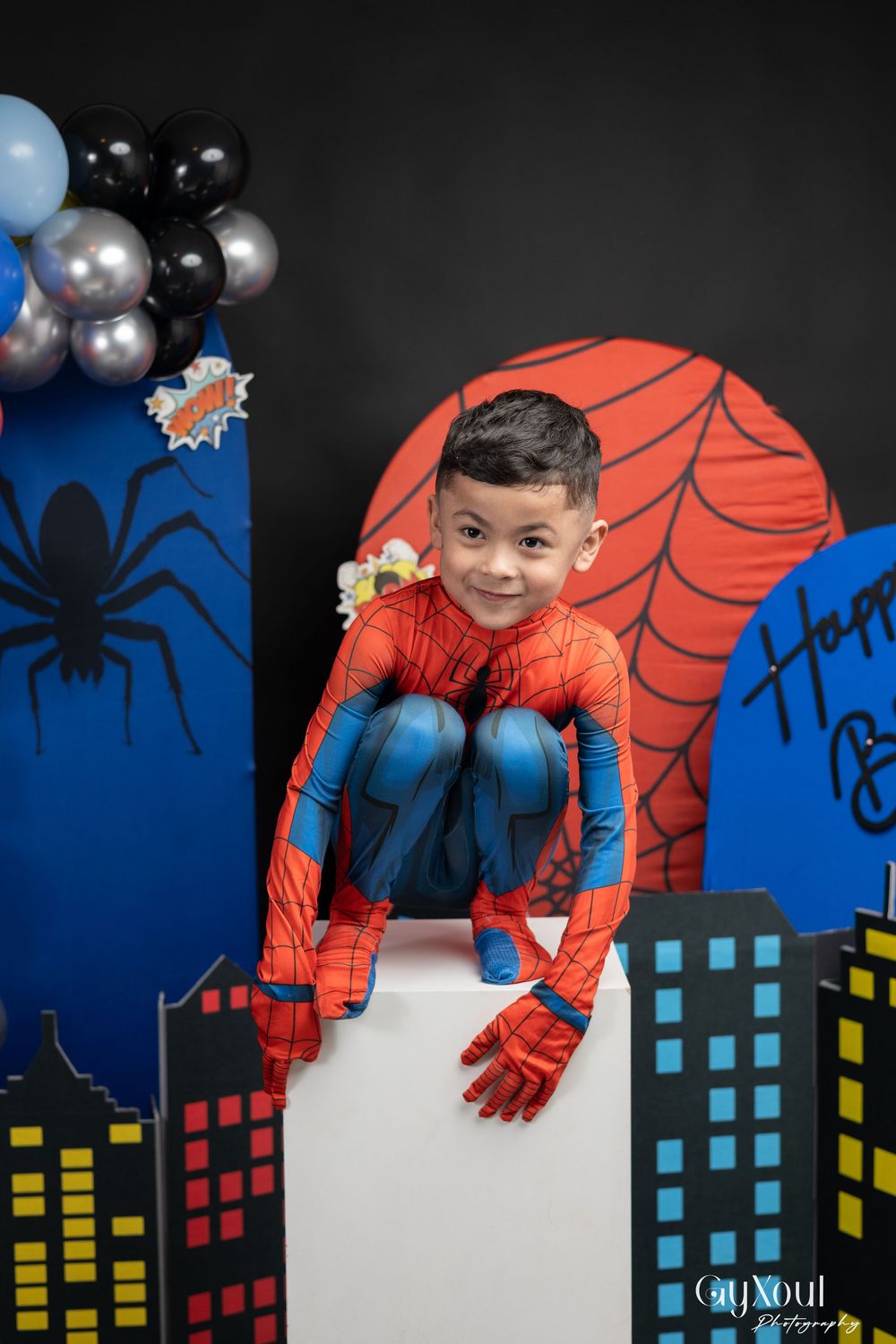 A child in a Spider-Man costume poses against a colorful cityscape backdrop with spiderweb decorations and silver balloons.