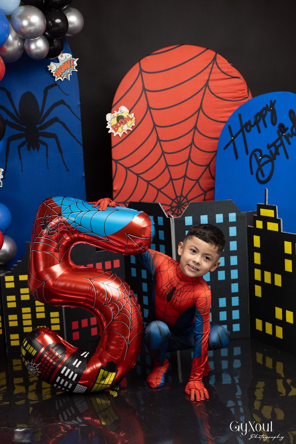 Spider-themed birthday party setup with red balloon number 5, web decorations, and city skyline backdrop against dark background.