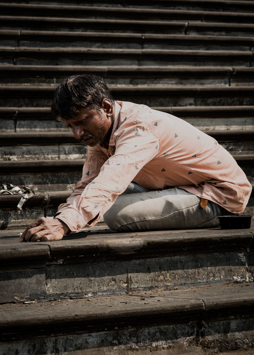 Man in pink shirt crouching on worn stone steps in natural sunlight.